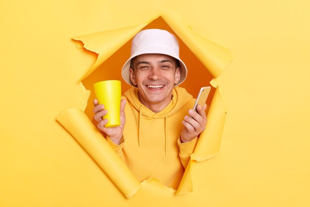 Portrait of extremely happy man wearing hoodie and panama posing in hole in yellow paper wall holding smart phone browsing internet and enjoying beverage from cup