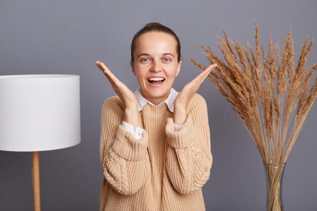 Portrait of extremely happy surprised woman wearing beige sweater standing against gray wall with lamp and dried flowers raised arms screaming happily hearing good news