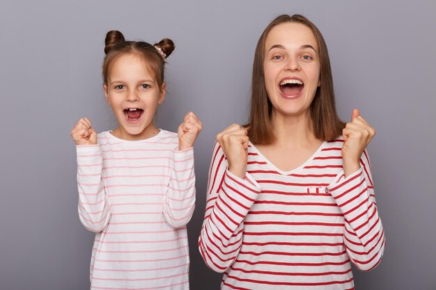 Portrait of extremely happy woman and little girl with hair buns wearing casual clothes clenching fists rejoicing winning standing isolated over gray background