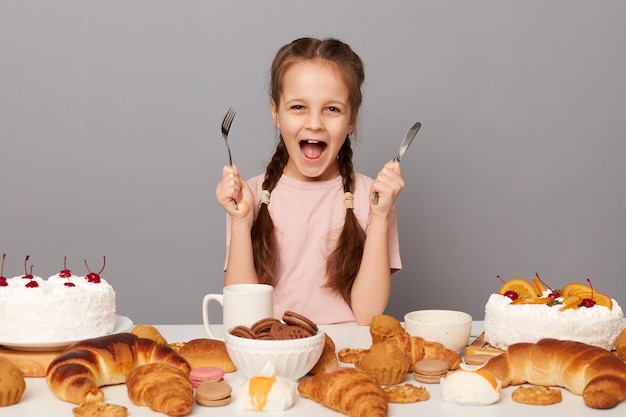 Portrait of extremely hungry funny female child with pigtails sitting at table with delicious confectionery isolated over gray background holding knife and fork yelling with crazy look