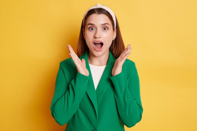Portrait of extremely surprised woman wearing green jacket and hair band raised arms keeps mouth widely opened being very shocked posing isolated over yellow background