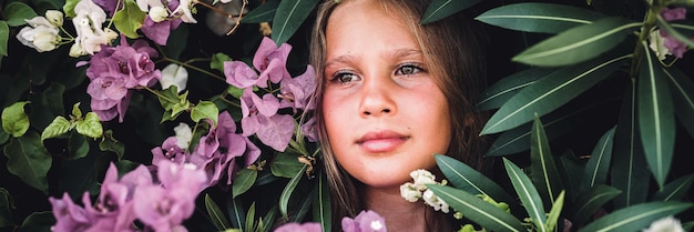 Portrait face of candid beautiful little kid girl of eight years old with brown eyes on background of green plants and pink flowers during a summer vacation travel gen z mental health concept banner