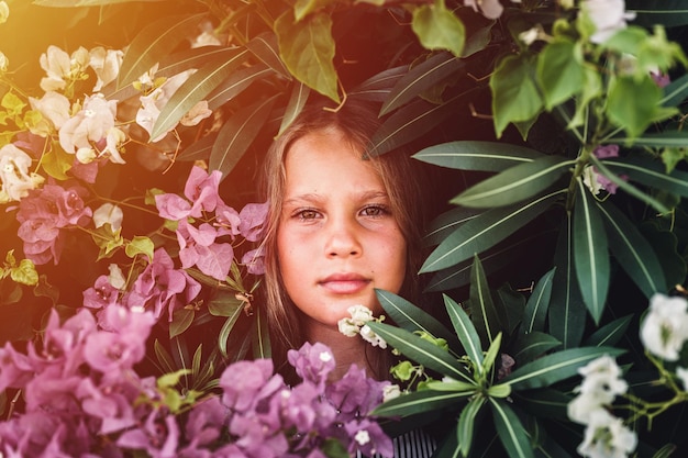 Portrait face of candid beautiful little kid girl of eight years old with brown eyes on background of green plants and pink flowers during a summer vacation travel gen z mental health concept flare