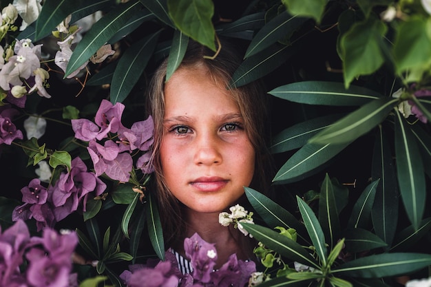 Portrait face of candid beautiful little kid girl of eight years old with green brown eyes on background of green plants and pink flowers during a summer vacation travel gen z mental health concept