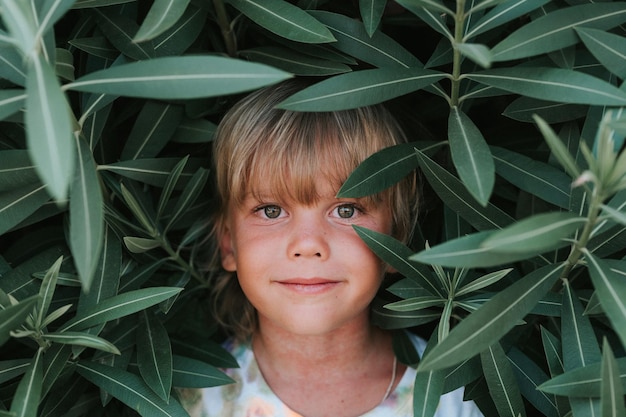 Portrait face of candid happy little kid boy of five years old with problem allergic skin and long blond hair and green eyes on background of green plants during a summer vacation travel