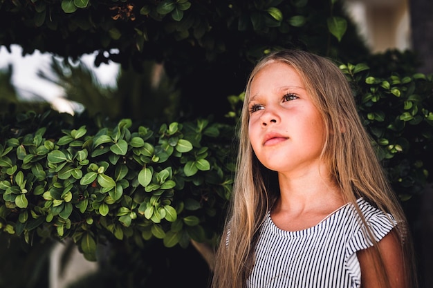 Portrait face of candid happy little kid girl of eight years\
old with long blond hair and green eyes on background of green\
plants during a summer vacation travel gen z mental health\
concept