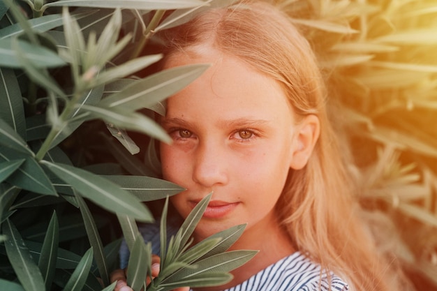 Portrait face of candid happy little kid girl of eight years old with long blond hair and green eyes on background of green plants during a summer vacation travel gen z mental health concept flare