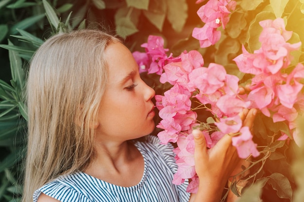 Portrait face of candid little kid girl of eight years old on background of green plants and admired enjoy smell pink flowers during a summer vacation travel gen z mental health concept flare