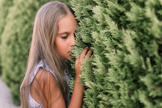 Portrait face of candid offended upset little kid girl of eight\
years old with long blond hair and green eyes on background of\
green plants during a summer vacation travel gen z mental health\
concept