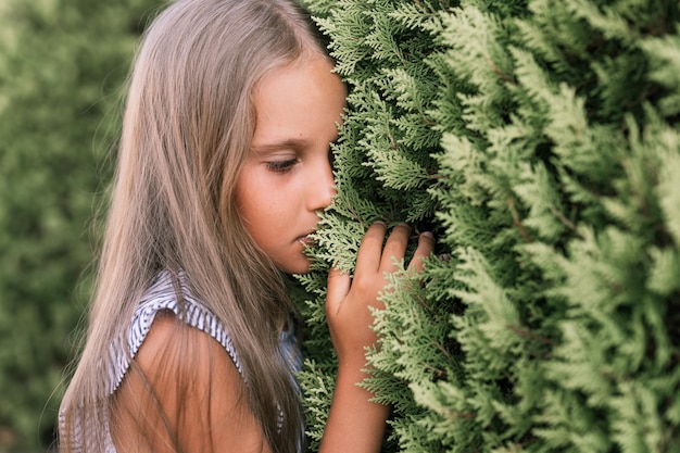 Portrait face of candid offended upset little kid girl of eight years old with long blond hair and green eyes on background of green plants during a summer vacation travel gen z mental health concept