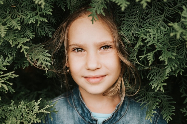 Portrait of the face of a cute happy caucasian candid healthy eight year old kid girl surrounded by branches and leaves of green plant thuja or cypress on nature outdoor