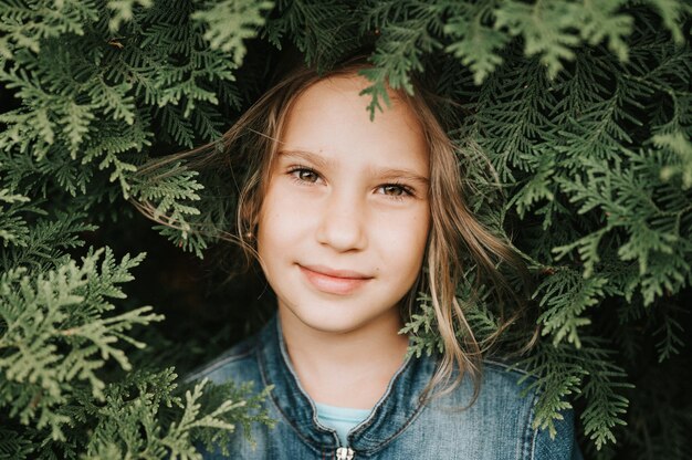 portrait of the face of a cute happy caucasian candid healthy eight year old kid girl surrounded by branches and leaves of green plant thuja or cypress on nature outdoor
