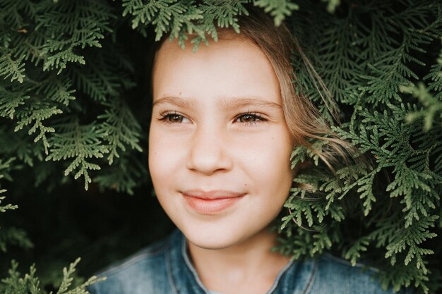 portrait of the face of a cute happy caucasian candid healthy eight year old kid girl surrounded by branches and leaves of green plant thuja or cypress on nature outdoor