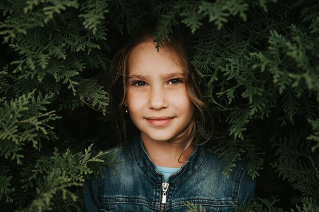 portrait of the face of a cute happy caucasian candid healthy eight year old kid girl surrounded by branches and leaves of green plant thuja or cypress on nature outdoor