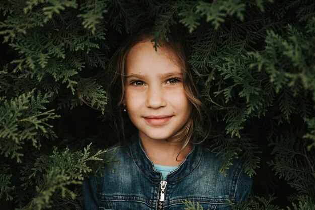 Portrait of the face of a cute happy caucasian candid healthy\
eight year old kid girl surrounded by branches and leaves of green\
plant thuja or cypress on nature outdoor