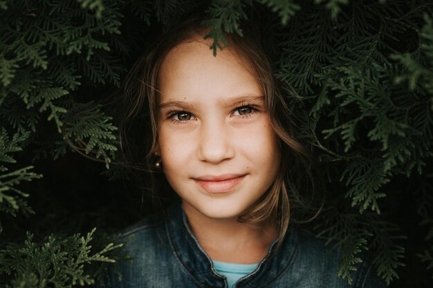 Portrait of the face of a cute happy caucasian candid healthy eight year old kid girl surrounded by branches and leaves of green plant thuja or cypress on nature outdoor