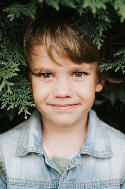 Portrait of the face of a cute little happy caucasian candid healthy five year old kid boy surrounded by branches and leaves of green plant thuja or cypress on nature outdoor