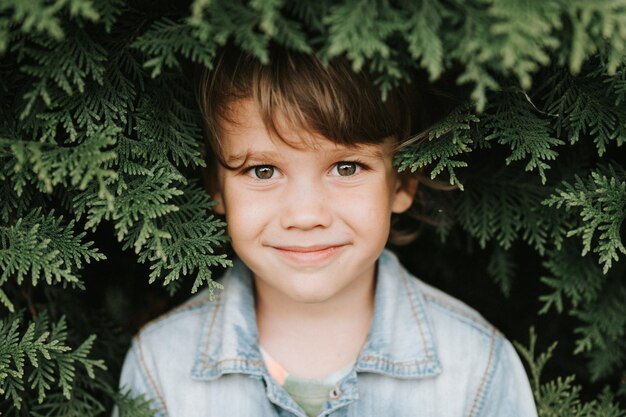 Portrait of the face of a cute little happy caucasian candid healthy five year old kid boy surrounded by branches and leaves of green plant thuja or cypress on nature outdoor