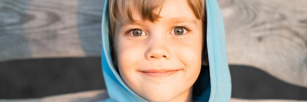日没のホームスクールと旅行バナーでゴールデンアワーの夏の自然の中で木の板の背景にフードの青いパーカーで幸せな笑顔の率直な5歳の子供の男の子の顔の肖像画