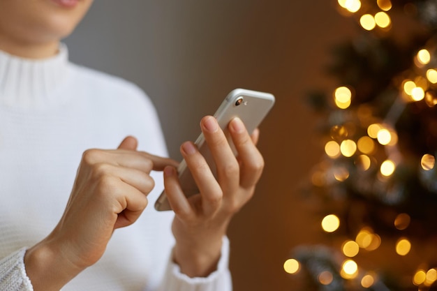 Portrait of faceless woman wearing white jumper posing near Christmas tree and holding smart phone checking social networks typing on cell phone using mobile phone for online communication