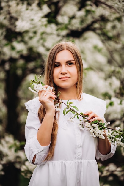 Un ritratto di una giovane donna bionda con un abito bianco come la neve si trova vicino a un melo in fiore. una ragazza cammina in un frutteto di mele in fiore in primavera