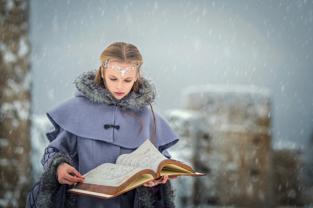 Portrait of a fairy-tale elf girl with a magic book in her hands against the backdrop of winter nature and magical fortress