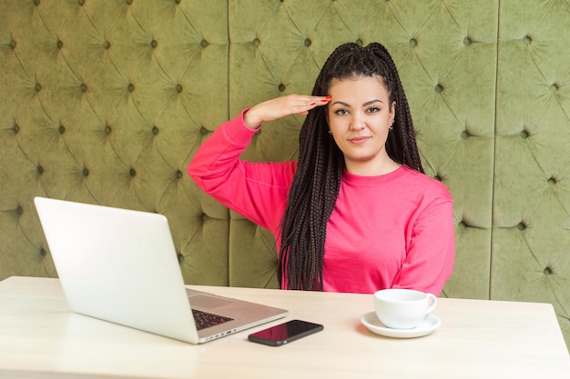 Portrait of faithful young girl freelancer with black dreadlocks hairstyle in pink blouse sitting in cafe and holding hand on head making salute looking at camera with serious face indoor