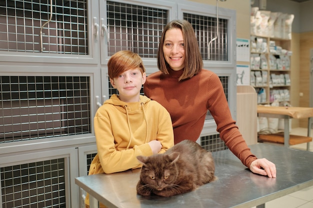 Photo portrait of family of two smiling at camera while standing at vet clinic with their cat on the table