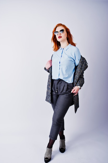 Portrait of a fantastic redheaded girl in blue shirt, grey overcoat posing with sunglasses in the studio.