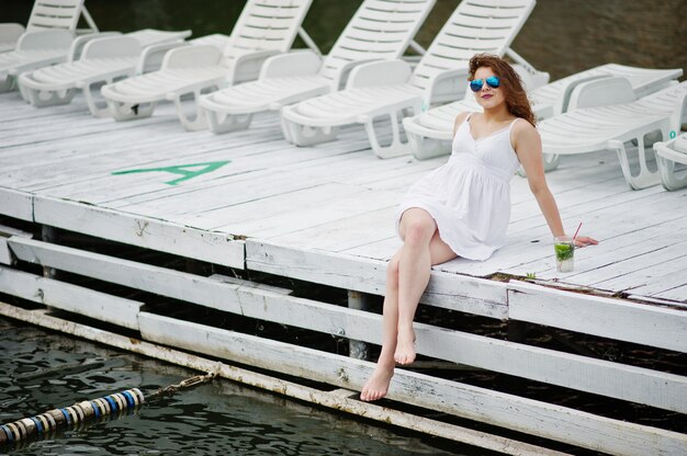 Portrait of a fantastic young girl with sunglasses and her drink sitting on white wooden harbour next to the water.