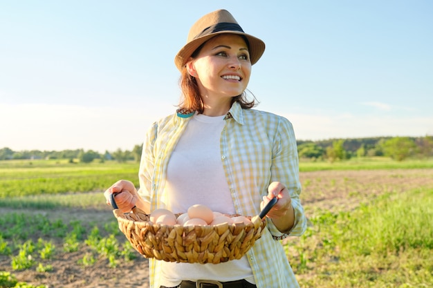 Portrait of farmer woman in hat with basket of fresh egg. Agriculture, farming organic eco products. Country road, countryside, sunset, nature, garden background