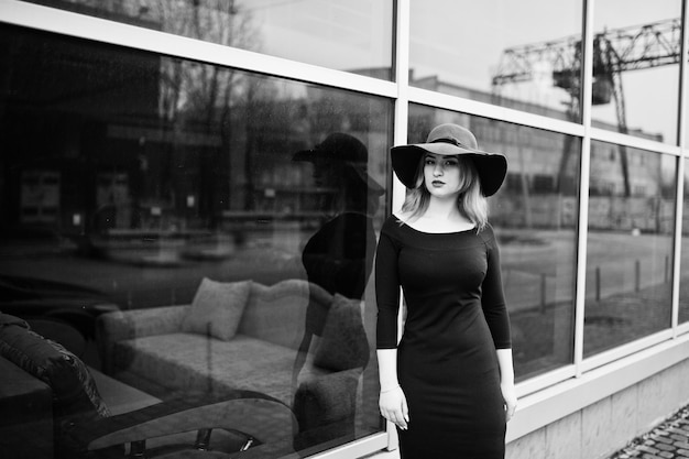 Photo portrait of fashion red haired girl on red hat and black dress with bright make up posed against large window photo toned style instagram filters black and white photo