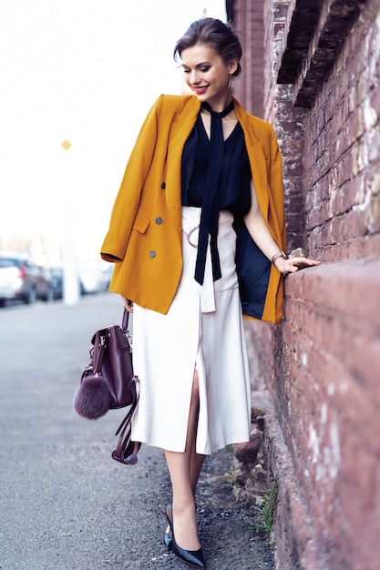 Portrait fashion woman walking on street . She wears yellow jacket, smiling to side.