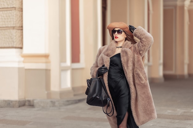 Portrait of fashion woman in a warm fur coat hat and sunglasses on a bright sunny day in the city