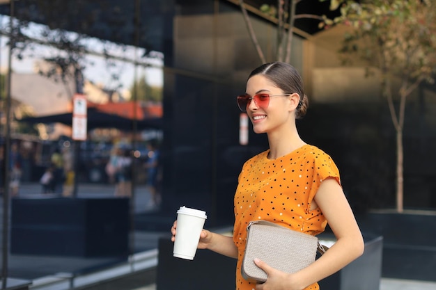 Portrait fashion woman in yellow dress holding coffee cup walking on street of the city smiling to side