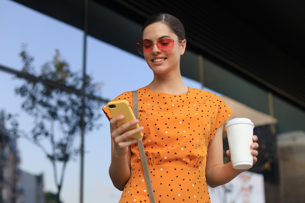 Portrait fashion woman in yellow dress in sunglasses walking on street and holding smartphone in her hand