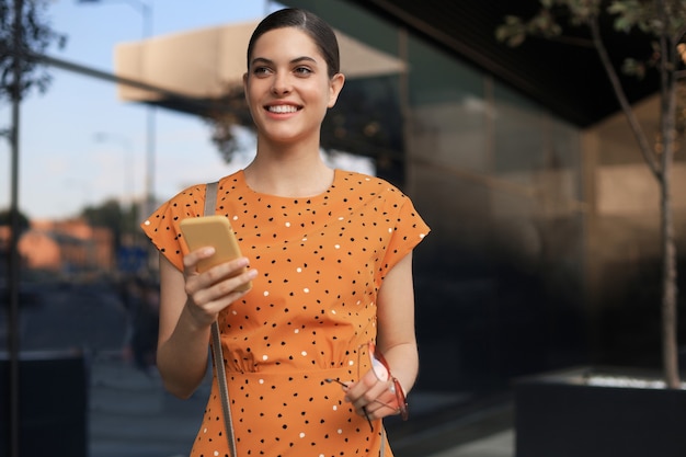 Портрет модной женщины в желтом платье, идущей по улице и держащей смартфон в руке.