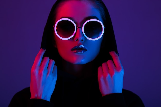 Portrait of fashion young girl in a black sweater with a hood and round sunglasses in red and blue neon light in the studio