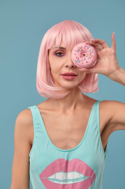 Portrait of a fashionable girl in a pink wig and with a pink donut in her hands keeps to the eye shows tongue