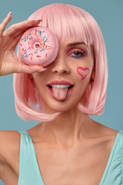 Portrait of a fashionable girl in a pink wig and with a pink donut in her hands keeps to the eye shows tongue