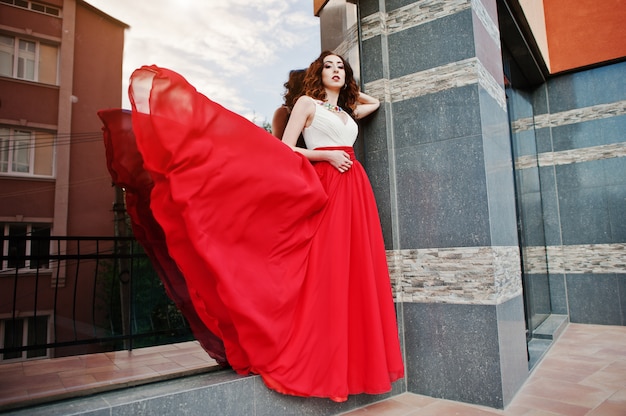 Portrait of fashionable girl at red evening dress posed background mirror window of modern building. Blowing dress in the air