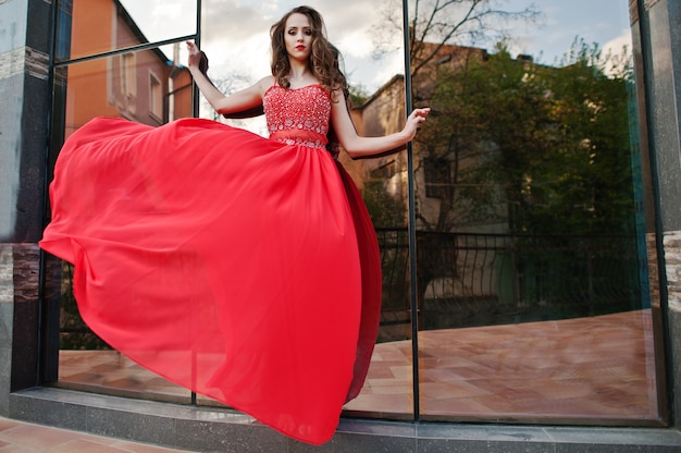 Portrait of fashionable girl at red evening dress posed background mirror window of modern building. Blowing dress in the air