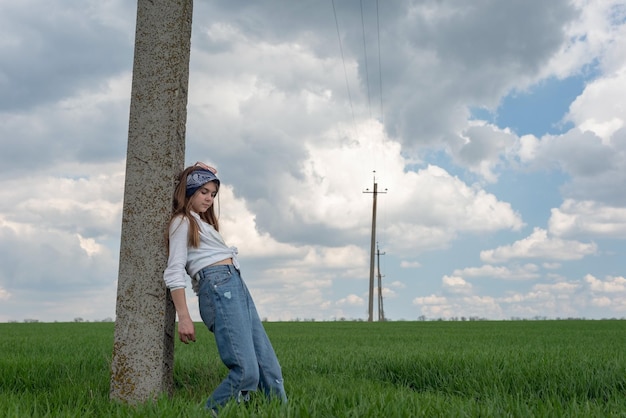 Ritratto di una piccola ragazza adolescente alla moda che posa da un pilastro in un paesaggio rurale del campo di erba verde in jeans