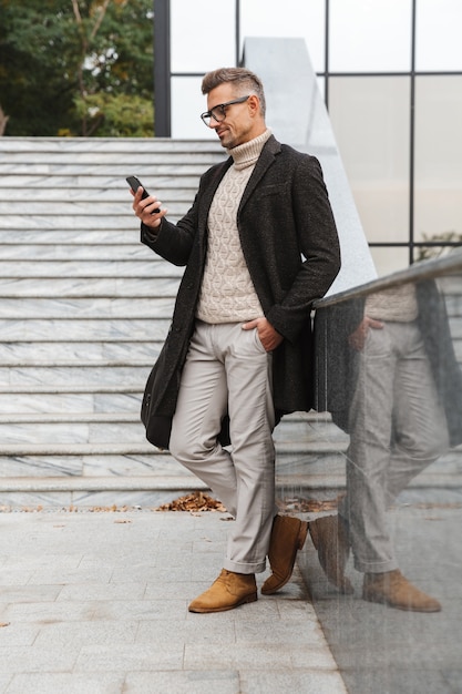 Portrait of fashionable man 30s wearing eyeglasses, walking through city street and using smartphone