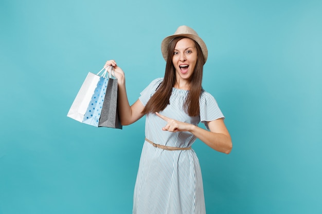 Portrait fashionable smiling beautiful caucasian woman in summer dress, straw hat holding packages bags with purchases after shopping isolated on blue pastel background. Copy space for advertisement.