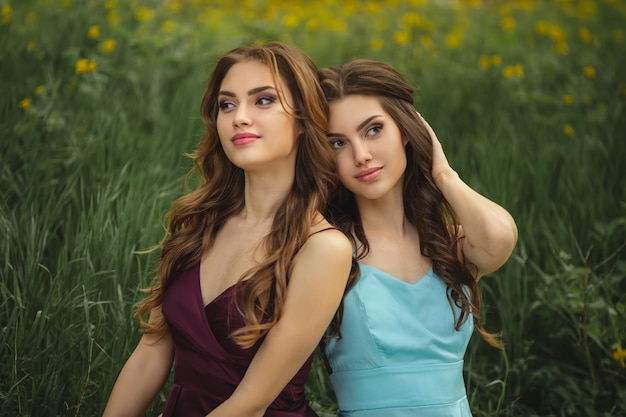 Portrait of fashionable twins models with perfect make-up and hairstyle sitting on green grass