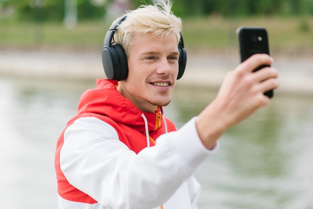 Portrait of fashionable young hipster man wearing trendy zipper hoodie having fun taking selfportrait with smart phone outdoors People lifestyle and technology concept