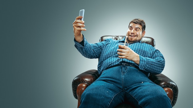 Portrait of fat caucasian man wearing jeanse and whirt sitting in a brown armchair on gradient grey background. Making selfie with the cola, laughting. Overweight, carefree. Concept of weight loss.