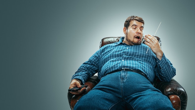 Portrait of fat caucasian man wearing jeanse and whirt sitting in a brown armchair isolated on gradient grey background. Emotional watching TV and changing channels, laughting. Overweight, carefree.