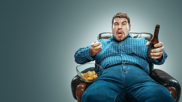 Portrait of fat caucasian man wearing jeanse and whirt sitting in a brown armchairbackground